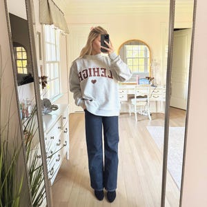 May include: A light gray sweatshirt with the word "LEHIGH" in maroon lettering and a maroon heart graphic. The sweatshirt is paired with dark blue jeans and black shoes. The image is taken in a well-lit room with a mirror.