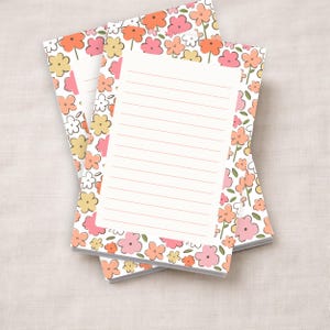 May include: Two floral-patterned notepads with a white lined interior. The covers feature a repeating pattern of pink, orange, yellow, and white flowers with green leaves on a white background. The notepads are stacked on top of each other.