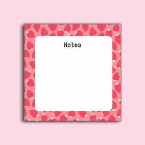 May include: A square notepad with a pink background and a pattern of red strawberries. The notepad has a white rectangular space in the center with the word "Notes" in black text.