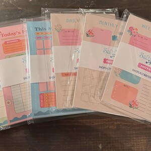 Op de afbeelding: Vijf rechthoekige papieren notitieblokken in doorzichtige plastic hoezen, elk met pastelkleurige lay-outs voor planning. De ontwerpen bevatten koppen zoals Today's Plan, Daily, Monthly en Weekly Planner, met secties voor takenlijsten en notities.