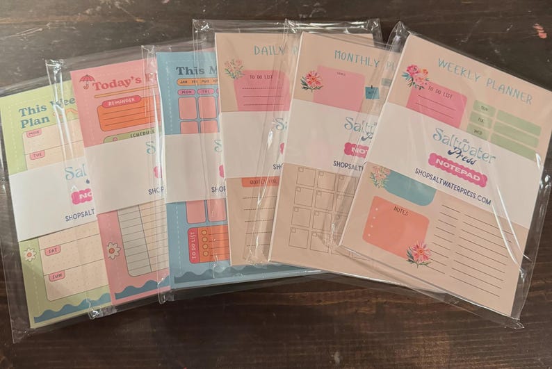 Op de afbeelding: Vijf rechthoekige papieren notitieblokken in doorzichtige plastic hoezen, elk met pastelkleurige lay-outs voor planning. De ontwerpen bevatten koppen zoals Today's Plan, Daily, Monthly en Weekly Planner, met secties voor takenlijsten en notities.
