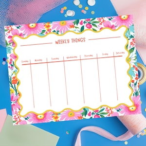 Puede incluir: Un planificador semanal floral con un interior blanco y un borde floral rosa, amarillo y verde. El planificador tiene las palabras "WEEKLY THINGS" en la parte superior y los días de la semana enumerados a continuación. El fondo es azul con elementos decorativos.