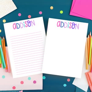 May include: Two white notepads with the name "ADDISON" in colorful letters. One notepad has lined paper, and the other is blank. Colored pencils and pink stationery are arranged around the notepads on a teal surface with colorful confetti.