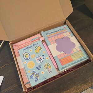 Puede incluir: Una caja de regalo con art&iacute;culos de papeler&iacute;a. Incluye un planificador, pegatinas, un bol&iacute;grafo y un bloc de notas. Las pegatinas tienen un tema de lim&oacute;n. El planificador y el bloc de notas tienen una combinaci&oacute;n de colores pastel con una nota adhesiva en forma de flor.
