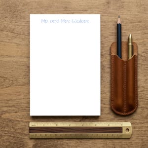 Peut inclure: Un bloc-notes rectangulaire blanc avec le texte Mr. and Mrs. Waters imprimé en bleu en haut, accompagné d'un crayon bleu foncé et d'un stylo doré dans un étui en cuir marron, placé au-dessus d'une règle en bois mesurant 20 cm de long.