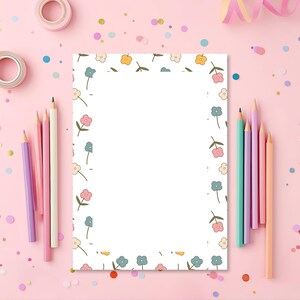 Könnte beinhalten: Ein weißes Blatt Papier mit einem floralen Rand in Pastellfarben, umgeben von Buntstiften, Konfetti und rosa Washi-Tape. Die Stifte sind in Rosa-, Lila- und Pfirsichtönen gehalten. Der Hintergrund ist hellrosa.