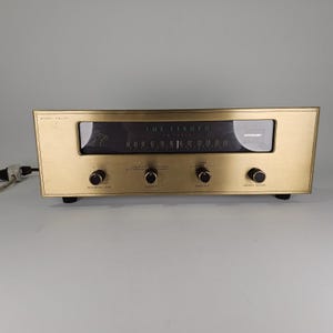 May include: A vintage The Fisher FM tuner with a brushed gold-tone metal faceplate. The device features a central black display panel with frequency markings, four black control knobs labeled recording level, selector, sensitivity, and station tuning.