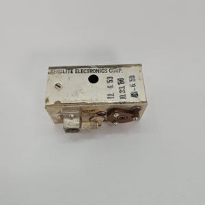 May include: A rectangular silver-toned metal electronic component by Aerolite Electronics Corp. The front face features a circular hole, a screw, and stamped dates including 11. 6. 53, 10. 23. 53, and 10. 6. 53. A small brown circular connector and a metal terminal are attached.