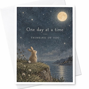 May include: A greeting card featuring a brown rabbit sitting on a grassy cliff overlooking a body of water under a starry night sky with a full moon. The text reads One day at a time and THINKING OF YOU in white lettering.