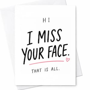 May include: A white greeting card with the text "HI I MISS YOUR FACE. THAT IS ALL." in black, handwritten-style font. A pink line underlines "YOUR FACE" with a small pink heart at the end. The card is inside a white envelope.