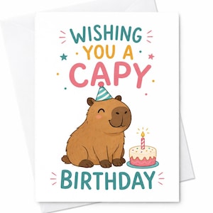 May include: A white greeting card featuring a brown cartoon capybara wearing a blue and white striped party hat. The text reads WISHING YOU A CAPY BIRTHDAY in colorful letters. A small pink and white birthday cake with a lit candle sits nearby.