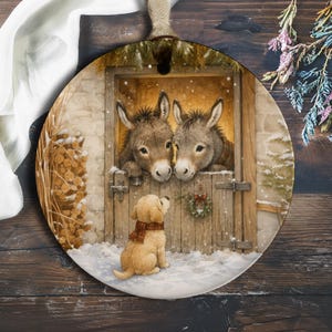 Puede incluir: Un adorno redondo de cerámica con una escena invernal de dos burros grises mirando desde una puerta de establo de madera, mientras un pequeño cachorro dorado con una bufanda a cuadros se sienta en la nieve mirando hacia arriba.