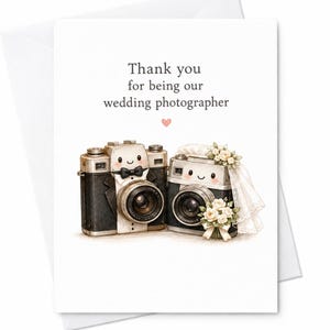 May include: A white greeting card featuring the text Thank you for being our wedding photographer with a small pink heart. Below, two illustrated cameras are depicted: one wearing a black tuxedo with a bowtie and one wearing a white bridal veil and a bouquet of white flowers.