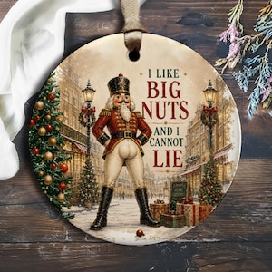 May include: A round ceramic ornament featuring a nutcracker in a red and white uniform standing in a snowy town square. The text reads I LIKE BIG NUTS AND I CANNOT LIE. The scene includes a decorated Christmas tree, street lamps, and gifts.