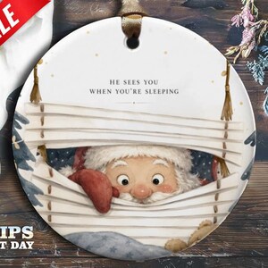 Könnte beinhalten: Ein rundes, weißes Keramikornament mit einer aufgedruckten Illustration vom Weihnachtsmann, der durch weiße horizontale Jalousien späht. Der Text HE SEES YOU WHEN YOU'RE SLEEPING ist in Schwarz über den Jalousien aufgedruckt. Ein braunes Band ist befestigt.