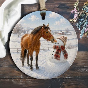 Puede incluir: Un adorno redondo de cerámica que muestra un caballo marrón con un cabestro rojo en un campo nevado junto a un muñeco de nieve. El muñeco de nieve lleva un gorro de punto estampado y una bufanda de cuadros rojos y marrones.