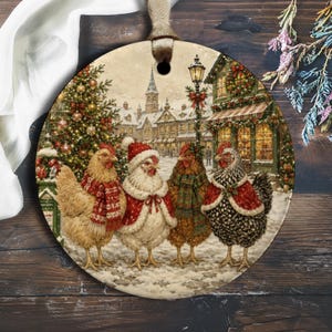 Puede incluir: Un adorno redondo de cerámica con una escena de pueblo nevado y cuatro pollos con accesorios de invierno. Un pollo lleva una bufanda roja, otro un gorro y capa de Papá Noel, otro una bufanda a cuadros y otro una capa roja con borde blanco en la nieve.