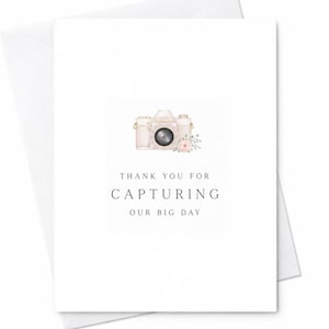 May include: A white greeting card featuring a watercolor illustration of a light pink camera adorned with a small floral accent. The text printed in black ink reads: THANK YOU FOR CAPTURING OUR BIG DAY. The card is paired with a matching white envelope.