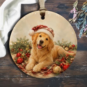 Könnte beinhalten: Ein rundes Keramikornament mit einem Golden Retriever, der eine rot-weiße Weihnachtsmannmütze trägt. Der Hund liegt auf einer karierten Decke, umgeben von Tannenzweigen, roten und goldenen Weihnachtskugeln sowie Tannenzapfen vor einem weichen Hintergrund.