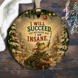 Könnte beinhalten: Ein rundes Keramikornament mit einer festlichen Winterszene, auf der ein Frosch mit Weihnachtsmütze und Schal auf einem Alligator reitet und ein Schwert hält. Der Text lautet: I WILL SUCCEED, BECAUSE I AM INSANE. Im Hintergrund sind verschneite Bäume.