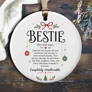 May include: A round white ceramic ornament featuring a red ribbon at the top. The text reads: BESTIE [bes-tee] noun. Someone who knows all your weirdness and chooses to stick around anyway. Your partner through the good days, bad days, laughter, tears, and every ridiculous moment in between. Completely irreplaceable. Decorated with red and green ornaments, a small red heart, and a green Christmas tree.