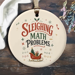 Pode incluir: Um ornamento redondo de cerâmica na cor creme com uma fita castanha. O design apresenta o texto "SLEIGHING MATH PROBLEMS" em vermelho, verde e preto, rodeado por símbolos matemáticos, flocos de neve, uma árvore de Natal e um trenó vermelho.