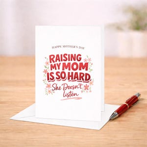 May include: A white Mother's Day greeting card with the text "Raising My Mom Is So Hard, She Doesn't Listen" in red and pink. A red pen rests on the wooden surface next to the card.