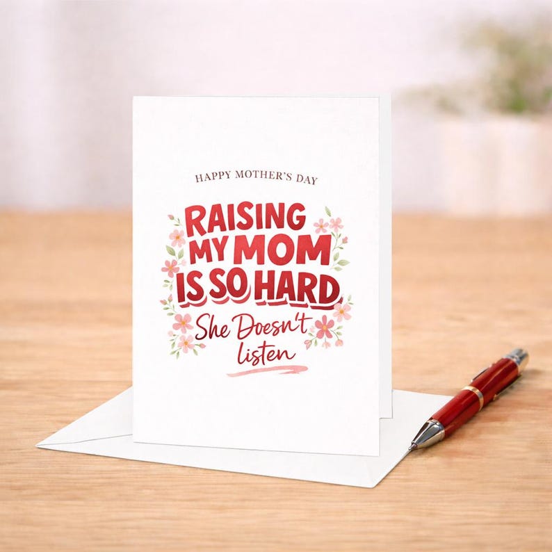May include: A white Mother's Day greeting card with the text "Raising My Mom Is So Hard, She Doesn't Listen" in red and pink. A red pen rests on the wooden surface next to the card.