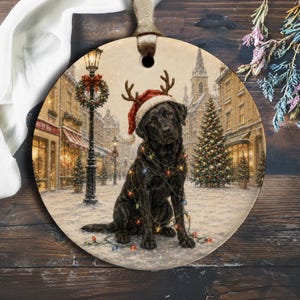 Könnte beinhalten: Ein rundes Keramikornament mit einem schwarzen Labrador, der eine rot-weiße Weihnachtsmütze mit Rentiergeweih trägt. Der Hund ist mit bunten Lichterketten umwickelt und befindet sich auf einem verschneiten Stadtplatz mit Weihnachtsbaum.