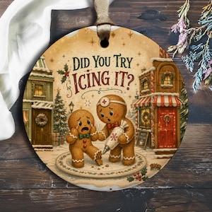 May include: A round ceramic ornament featuring a gingerbread person with a broken leg being treated by another gingerbread person holding an icing bag. The text reads DID YOU TRY ICING IT? The scene is set in a snowy village with festive buildings.