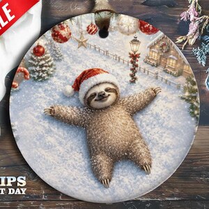 May include: A round ceramic ornament featuring a brown sloth wearing a red and white Santa hat, lying in the snow with arms and legs spread out. The background shows a winter scene with a decorated house, a street lamp, and hanging ornaments.