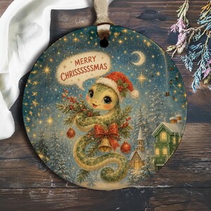 Op de afbeelding: Een rond keramieken ornament met een groene slang in een kerstmuts, die een dennentak met rode kerstballen en een gouden bel vasthoudt. De achtergrond toont een besneeuwd winterlandschap met een huis. De tekst luidt MERRY CHRISSSSSMAS.