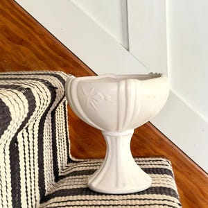 May include: A white, pedestal-style vase with a fluted body and a scalloped rim. The vase has a floral design and stands on a round base. The vase is approximately 8 inches tall and is placed on a black and white striped surface.