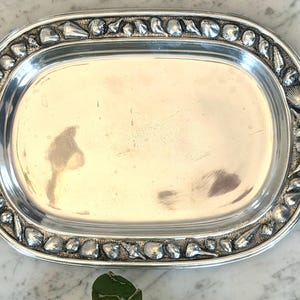 May include: A silver-colored, oval serving tray with a decorative border of seashells. The tray has a polished, reflective surface and a detailed edge. The design evokes a coastal or nautical theme, suitable for serving food or displaying decorative items.