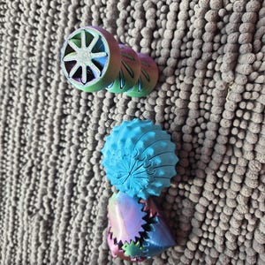 May include: A collection of colorful, 3D-printed geometric shapes. The shapes include a wheel-like design with a star pattern, a spiky blue sphere, and a cone-shaped piece with a jagged edge. The objects have a rainbow gradient color scheme.