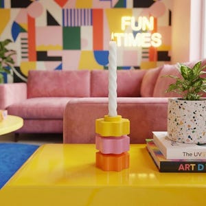 Peut inclure: Une bougie blanche torsadée est placée sur un bougeoir coloré empilé. Le bougeoir est composé de couches jaunes, roses et oranges en forme d'étoile. L'arrière-plan présente un canapé rose, un mur coloré et une enseigne au néon qui dit "FUN TIMES."