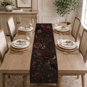 May include: A dark-toned table runner featuring a detailed print of deep red roses, winding vines, golden crescent moons, a vintage skeleton key, and black birds. The runner is centered on a light wood dining table with place settings.