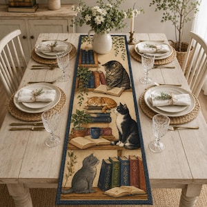 May include: A rectangular table runner featuring a printed illustration of multiple cats resting among stacks of books and potted plants. The design includes various cats in shades of grey, orange, and black and white, set against a cream background.