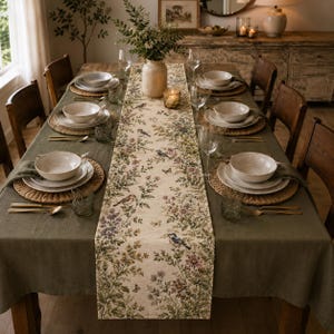 May include: A dining table set with a dark olive green tablecloth and a cream-coloured table runner featuring a botanical print of birds, butterflies, and flowers. The table is arranged with woven placemats, white ceramic bowls, plates, and gold cutlery.