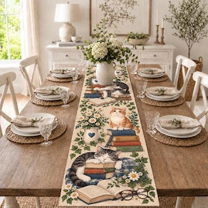 May include: A rectangular table runner featuring a printed design of cats resting on stacks of books, surrounded by green ivy vines and white daisy flowers. The runner is centered on a wooden dining table set with plates, napkins, and glasses.