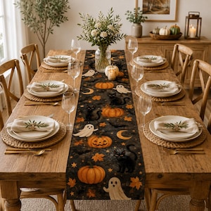 Puede incluir: Una mesa de comedor de madera preparada para seis personas con un camino de mesa oscuro con temática de Halloween que presenta calabazas, gatos negros, fantasmas y lunas crecientes. Cada cubierto incluye un mantel individual trenzado, un plato blanco, una servilleta doblada con vegetación y un vaso.