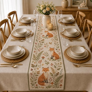 May include: A beige linen table runner featuring a repeating pattern of orange foxes, green ferns, and small mushrooms, centred on a dining table set with white ceramic bowls, woven placemats, clear glassware, and a vase of white flowers.