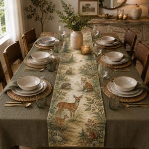 May include: A dining table set with a long, rectangular table runner featuring a printed forest scene with a deer, fawn, fox, hedgehog, rabbit, and squirrel. The table is arranged with white ceramic bowls, plates, woven placemats, and glassware.