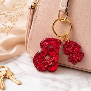 May include: A gold-toned metal keyring featuring two red resin charms with embedded glitter. One charm is shaped like the letter B, and the other is shaped like a hamsa hand. The keyring is attached to a light pink handbag, and two brass keys lie nearby.