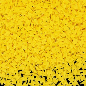 Könnte beinhalten: Nahaufnahme von gelben, blitzförmigen Streuseln. Die Streusel sind verstreut und erzeugen ein strukturiertes Muster. Die leuchtend gelbe Farbe kontrastiert mit dem dunklen Hintergrund und hebt die Formen hervor. Ideal zum Dekorieren von Kuchen und Gebäck.
