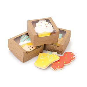 Puede incluir: Tres cajas de cartón marrón con ventanas de plástico transparente, cada una contiene una galleta decorada. Las galletas son una magdalena, un hombre de jengibre y una campana. Las cajas tienen un patrón de lunares dorados.