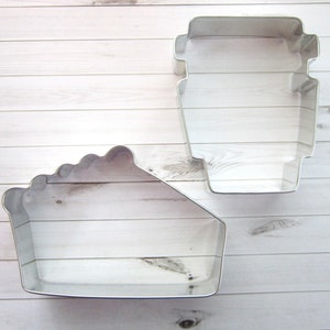 May include: Two metal cookie cutters, one shaped like a slice of pie and the other shaped like a coffee cup. Both cutters are silver in color and are on a white wooden background.