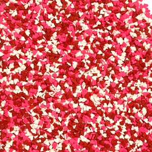 May include: A close-up view of a pile of heart-shaped sprinkles in shades of red, pink, and white. These small, decorative sprinkles are scattered, creating a textured surface. Ideal for decorating baked goods.