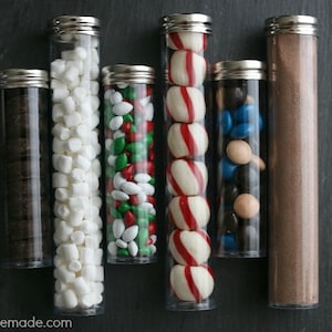 Op de afbeelding: Zes doorzichtige glazen buizen met zilveren deksels gevuld met verschillende snoepjes en chocolade. De buizen bevatten mini marshmallows, groene en rode snoepjes, pepermuntjes, chocolade snoepjes en chocoladepoeder.