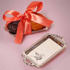 May include: Two small silver trays with ornate handles. One tray has a small orange and brown cookie on it, tied with a bright orange ribbon. The other tray has a silver bird design.